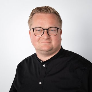 Nahaufnahme eines selbstbewussten Mannes mit Brille Ein Mann mit blonden Haaren und Brille lächelt freundlich vor einem hellen Hintergrund, gekleidet in ein schwarzes Hemd.