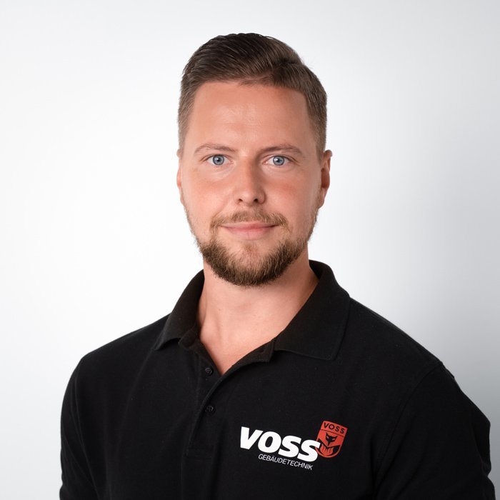Ein Mann mit einem gepflegten Bart trägt ein schwarzes Poloshirt mit dem Logo von VOSS Gebäude-Technik.
