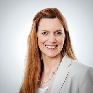 Professionelles Porträt einer lächelnden Frau mit langen Haaren Eine Frau mit langen, blonden Haaren lächelt freundlich in die Kamera, gekleidet in einem hellen Blazer und einer Perlenkette.