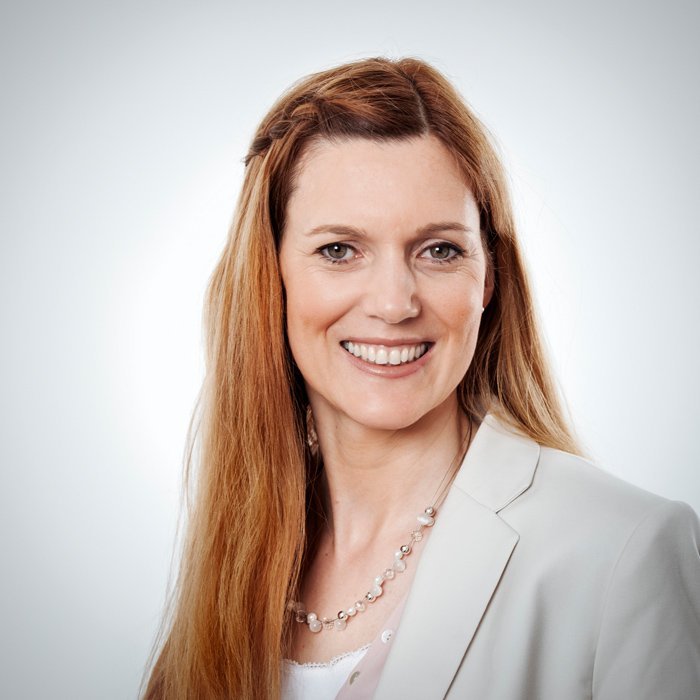 Eine Frau mit langen, blonden Haaren lächelt freundlich in die Kamera, gekleidet in einem hellen Blazer und einer Perlenkette.