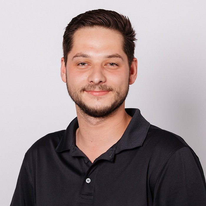 Ein junger Mann mit einem freundlichen Lächeln trägt ein schwarzes Polohemd und hat einen gepflegten Bart.