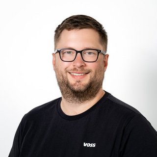 Lächelnder Mann mit Brille vor hellem Hintergrund Ein lächelnder Mann mit Brille trägt ein schwarzes T-Shirt mit dem Aufdruck 'VOSS' vor einem weißen Hintergrund.