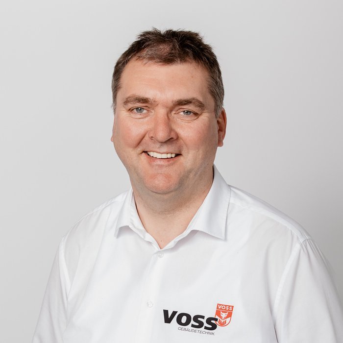 Ein lächelnder Mann trägt ein weißes Hemd mit dem Logo von VOSS Gebäudetechnik, vor einem neutralen Hintergrund.