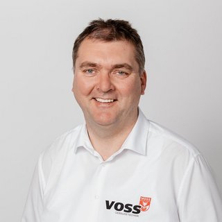 Lächelnder Mann in VOSS-Gebäudetechnik Hemd Ein lächelnder Mann trägt ein weißes Hemd mit dem Logo von VOSS Gebäudetechnik, vor einem neutralen Hintergrund.
