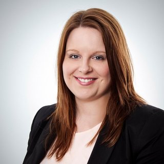 Freundliches Gesicht einer Geschäftsfrau mit langen Haaren Eine lächelnde Frau mit langen, braunen Haaren trägt einen schwarzen Blazer und ein helles Oberteil vor einem neutralen Hintergrund.
