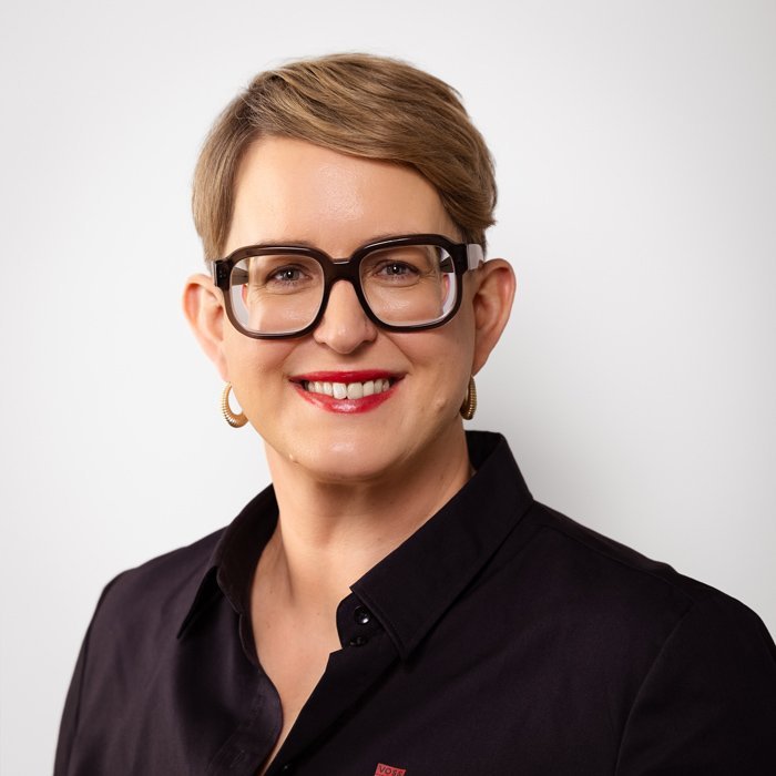 Eine lächelnde Frau mit kurzen, blonden Haaren trägt eine schwarze Bluse und große, modische Brillen.