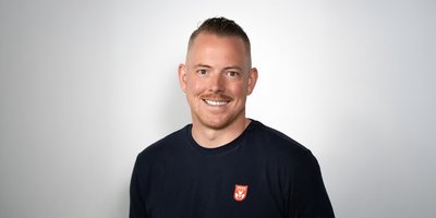 Ein lächelnder Mann mit kurzem Haar trägt ein schwarzes T-Shirt mit einem roten Logo auf der linken Brustseite.