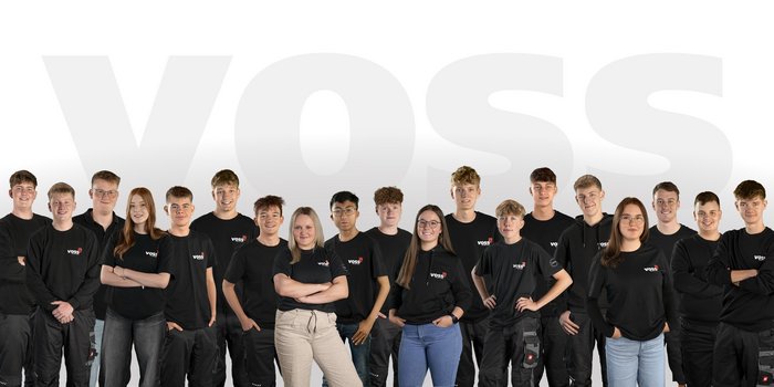 Eine Gruppe junger Menschen in schwarzen T-Shirts mit dem Logo 'VOSS' steht lächelnd vor einem hellen Hintergrund.
